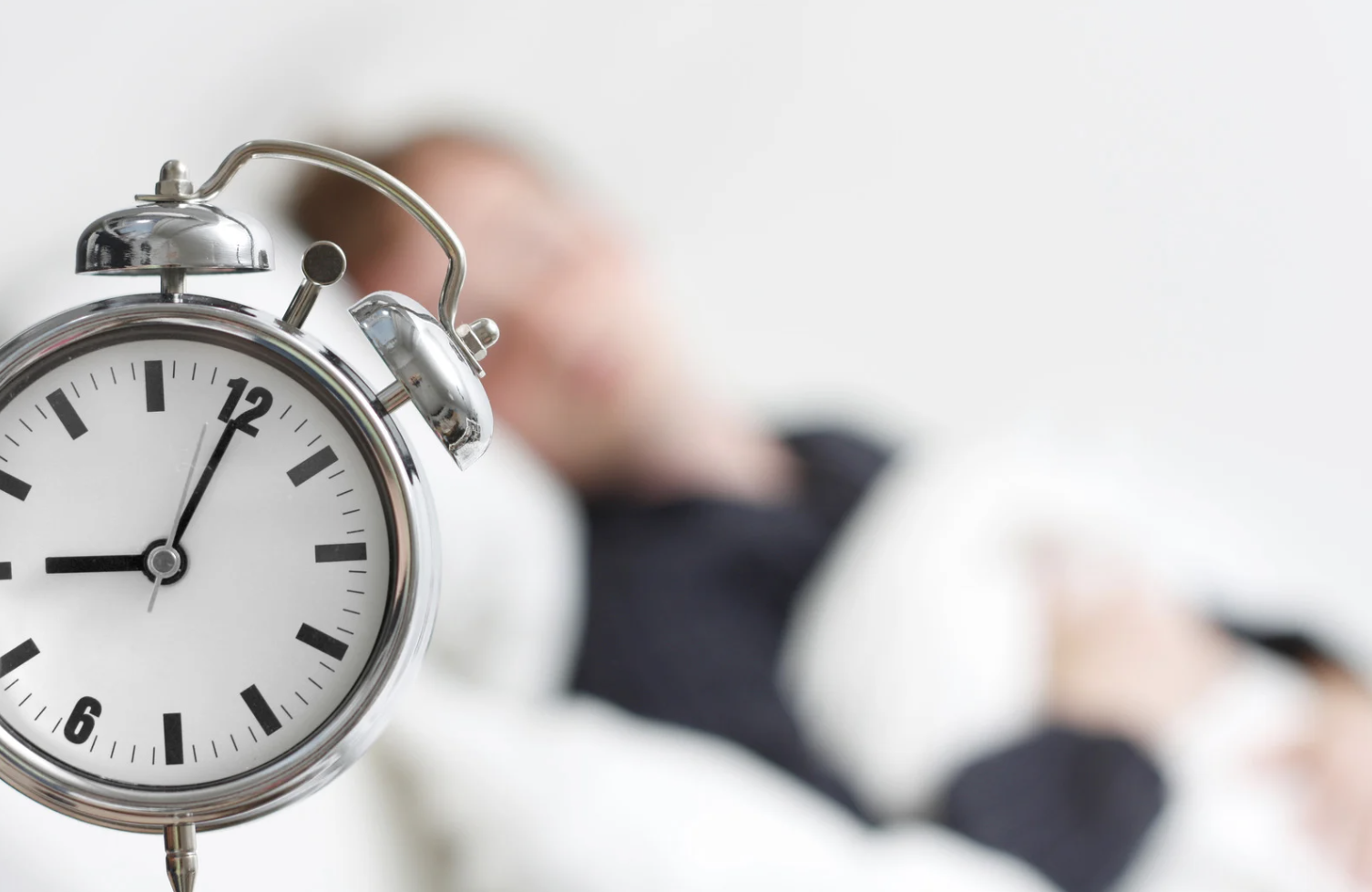 Close-up of an alarm clock showing 7:00 with a person sleeping in the background, representing rest and circadian rhythm.