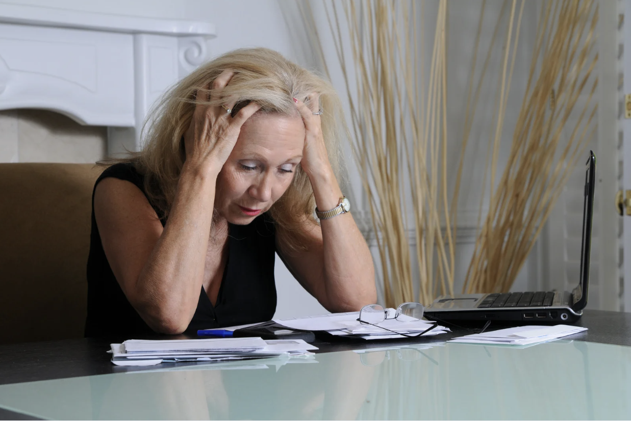 Older woman sitting at a desk with her hands on her head, looking stressed while surrounded by papers and a laptop.