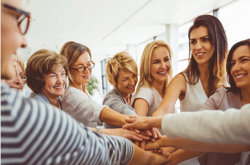 Group of smiling women standing together with their hands stacked in the center, showing unity, teamwork, and support in a bright indoor setting.