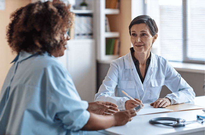 Female doctor in a white coat sitting at a desk, smiling and talking with a patient during a medical consultation.