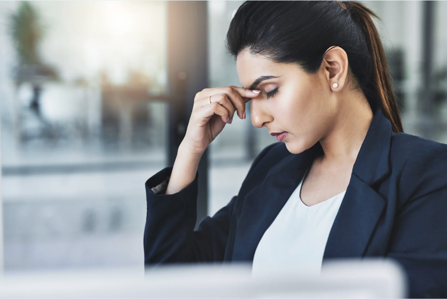 Woman in business attire sitting at a desk with eyes closed, holding her forehead in frustration or fatigue, representing stress or headache.
